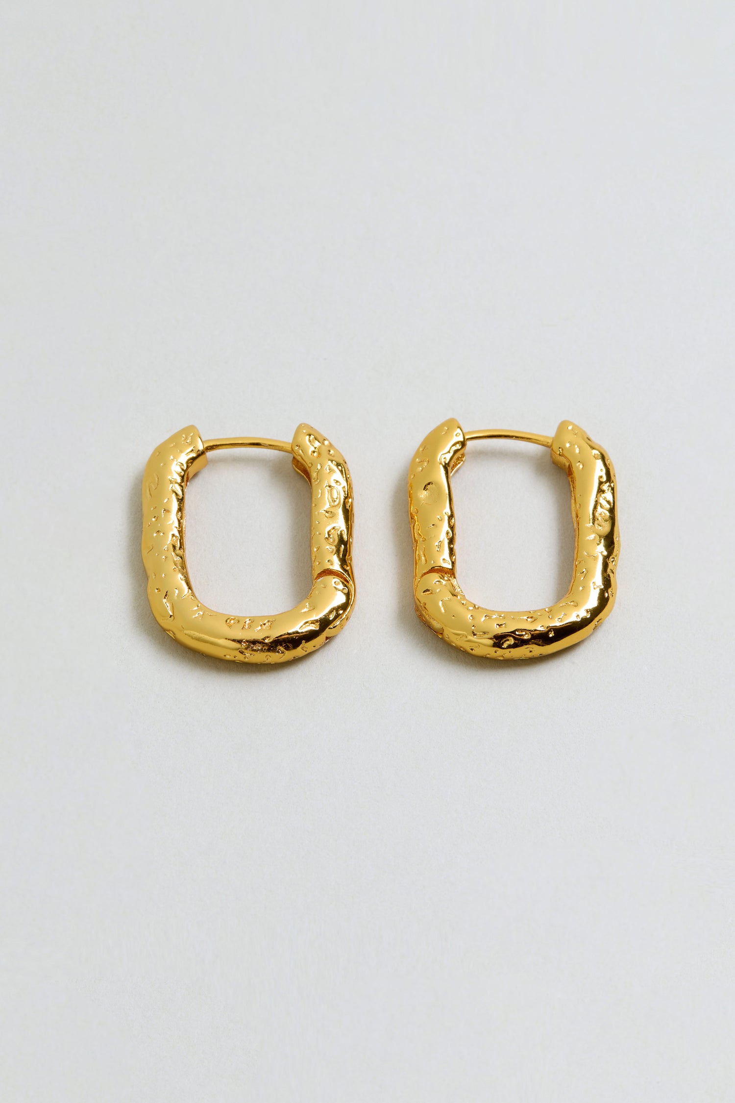 A pair of gold plated, rectangular hammered hoop earrings with a textured, uneven surface, displayed on a plain light background.