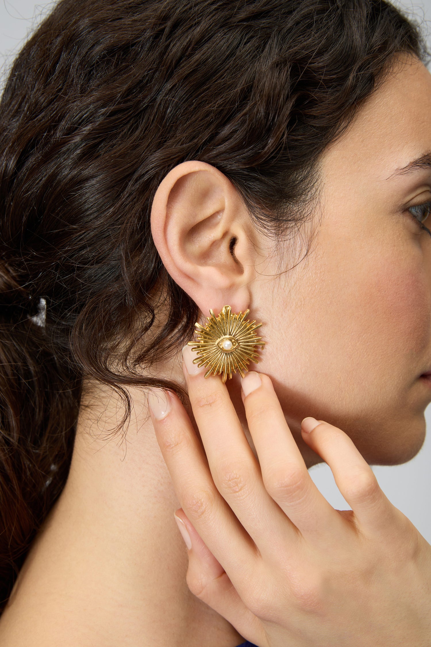 A person with dark, wavy hair wears Gold Plated Luna Pearl Earrings, featuring a sunburst design and pearl center, while gently touching their ear.