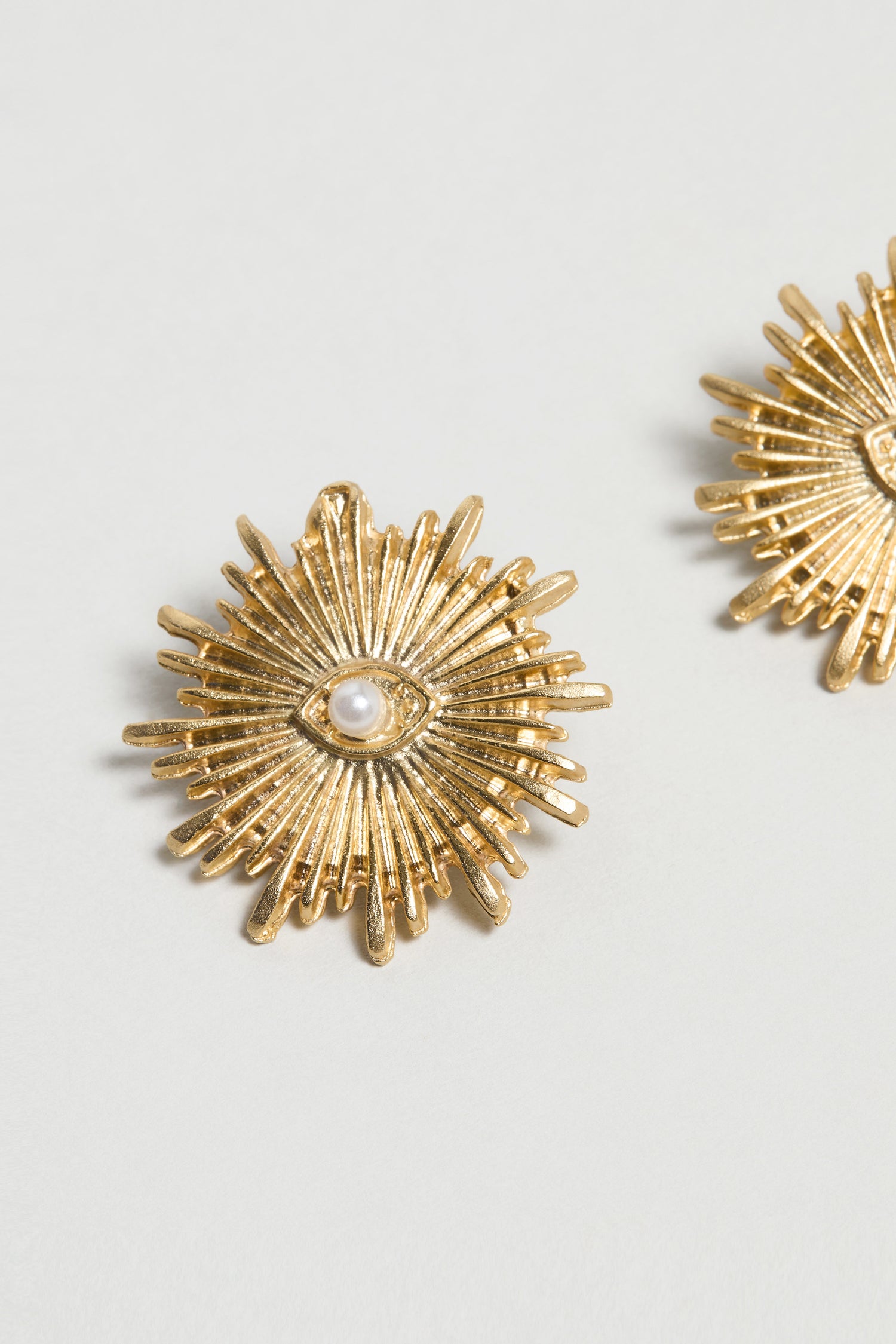 Gold Plated Luna Pearl Earrings, featuring a sunburst design and a striking pearl centre, are shown on a light background with part of the matching earring visible at the right edge.