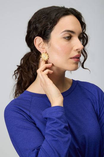 A woman with dark curly hair in a ponytail wears a blue top and touches striking Gold Plated Luna Pearl Earrings, featuring a sunburst design and luminous pearl center.