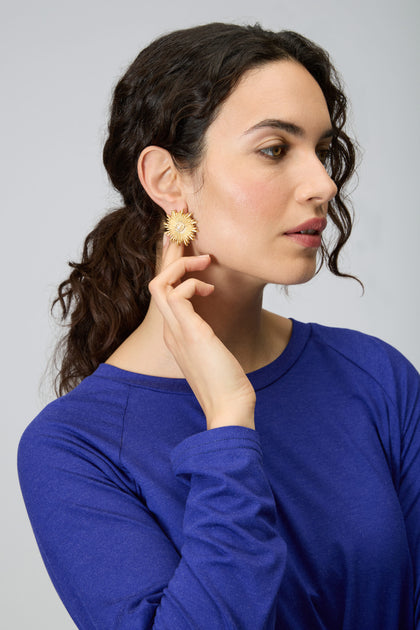 A woman with dark curly hair in a ponytail wears a blue top and touches striking Gold Plated Luna Pearl Earrings, featuring a sunburst design and luminous pearl center.