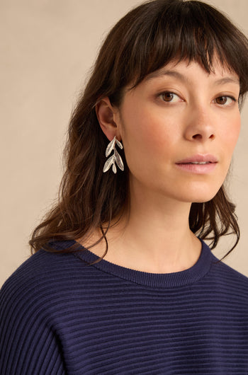 Woman with medium-length brown hair wearing a navy ribbed sweater and elegant silver plated leaf drop earrings, looking slightly past the camera against a neutral background.