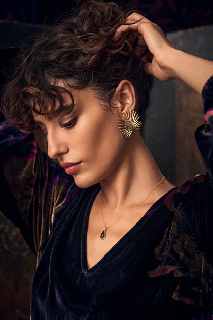 A person wears large, gold, fan-shaped Sunburst Fan Earrings and a gold necklace with a dark teardrop pendant.