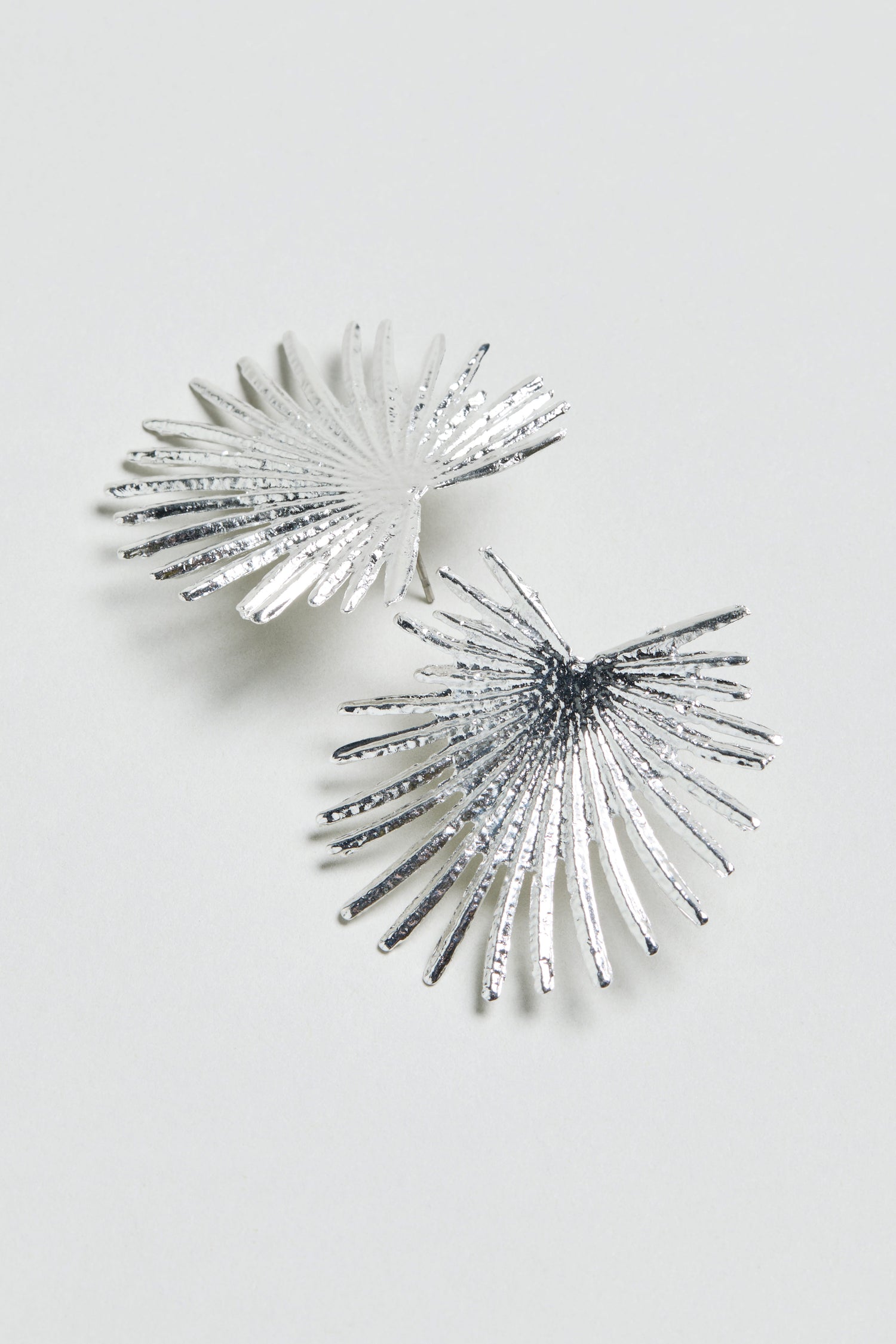 A pair of Sunburst Fan Earrings in silver metallic, featuring an abstract palm leaf design, displayed on a plain light gray background.