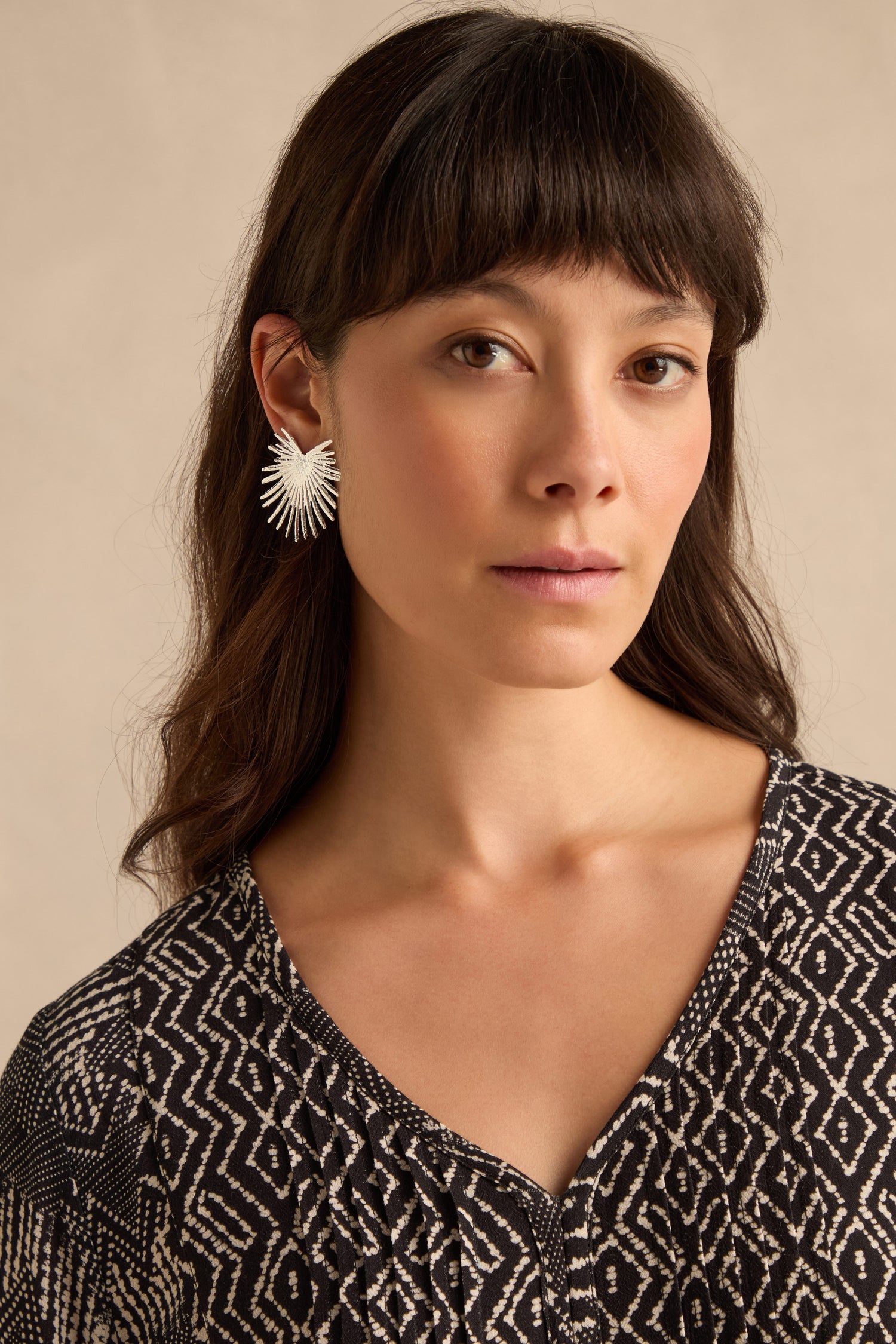 A woman with long dark hair and bangs wears a patterned black-and-white top and the Sunburst Fan Earrings, standing against a neutral background.