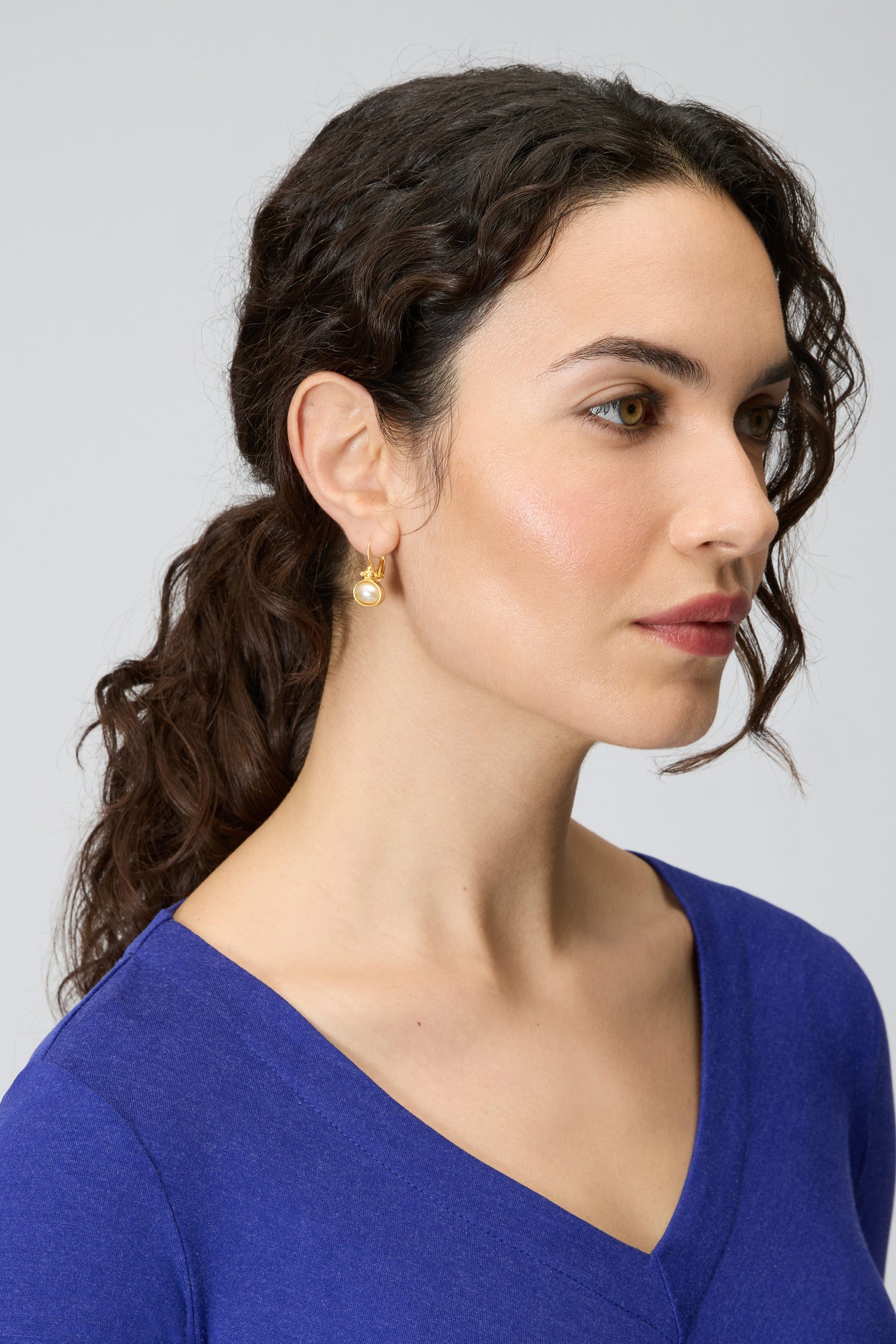 A woman with wavy brown hair in a low ponytail wears Gold Plated Pearl Drop Earrings and a blue V-neck top, looking to the right against a plain background.