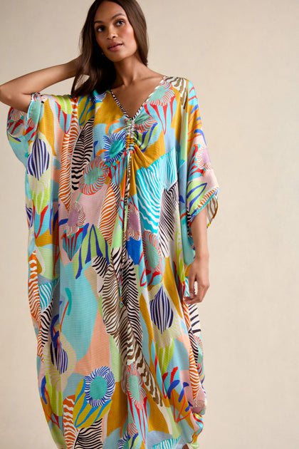 Woman wearing the Tropical Fantasy Kaftan, an oversized maxi dress with abstract, multicolored patterns, poses with one hand touching her hair against a plain background.