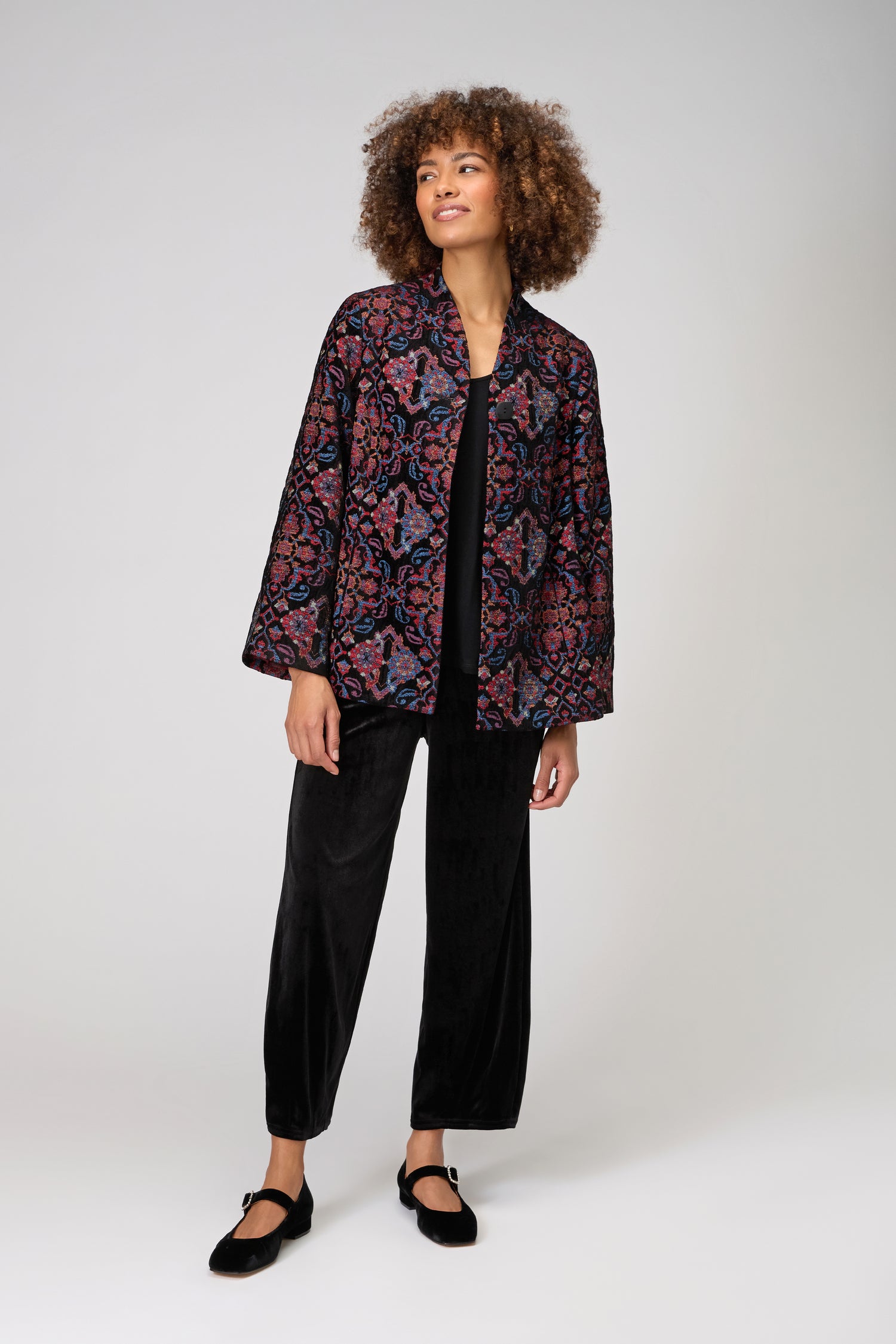 A person stands against a plain, light background wearing the Baroque Chenille Jacket over a black top and black pants.