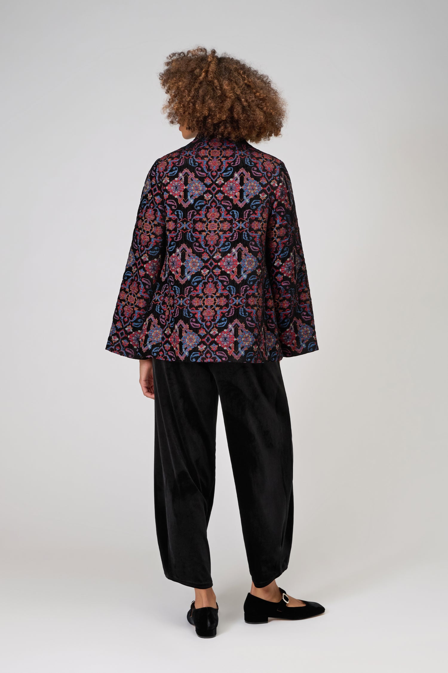 A person with curly hair, seen from behind, wears the luxurious Baroque Chenille Jacket over loose black pants and black loafers, standing against a plain light background.