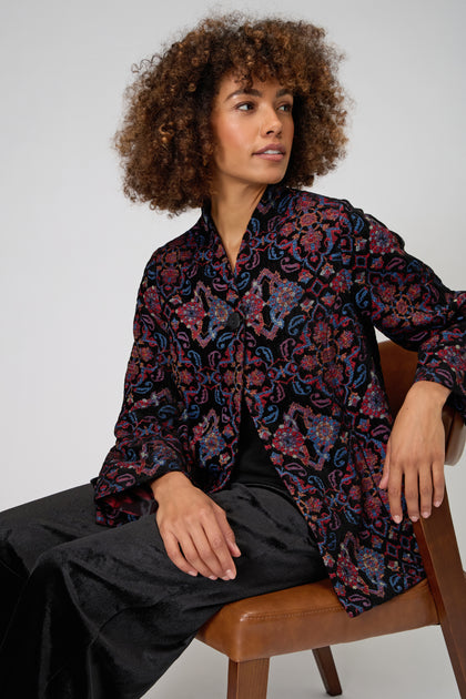 A woman with curly hair sits on a brown chair, wearing the Baroque Chenille Jacket and black pants, gazing slightly to the side against a plain background.