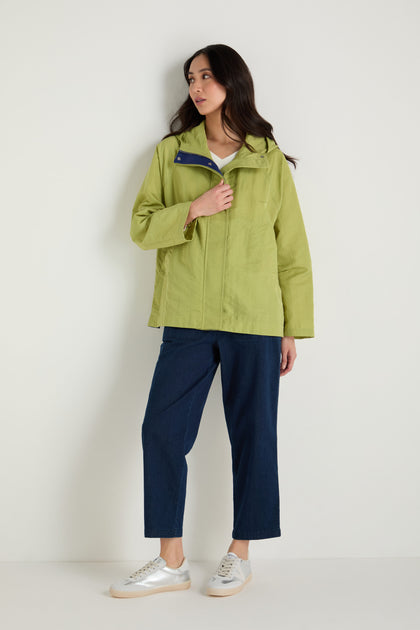 A woman stands against a plain wall wearing a light green Showerproof Boxy Rain Jacket, dark blue cropped pants, and white sneakers.