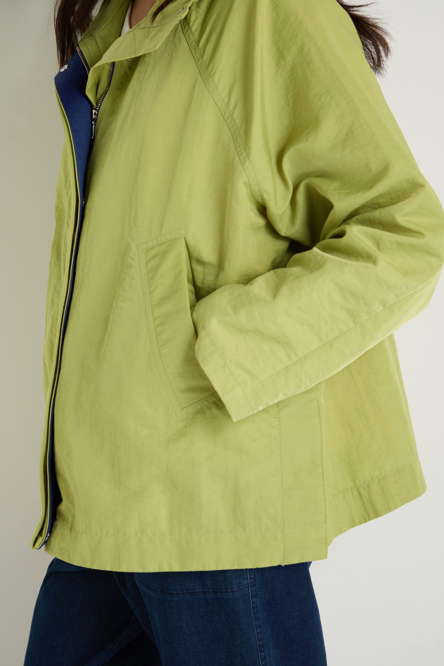 A person wearing the Showerproof Boxy Rain Jacket in light green with their left hand in the pocket, paired with dark blue pants, stands against a plain background.