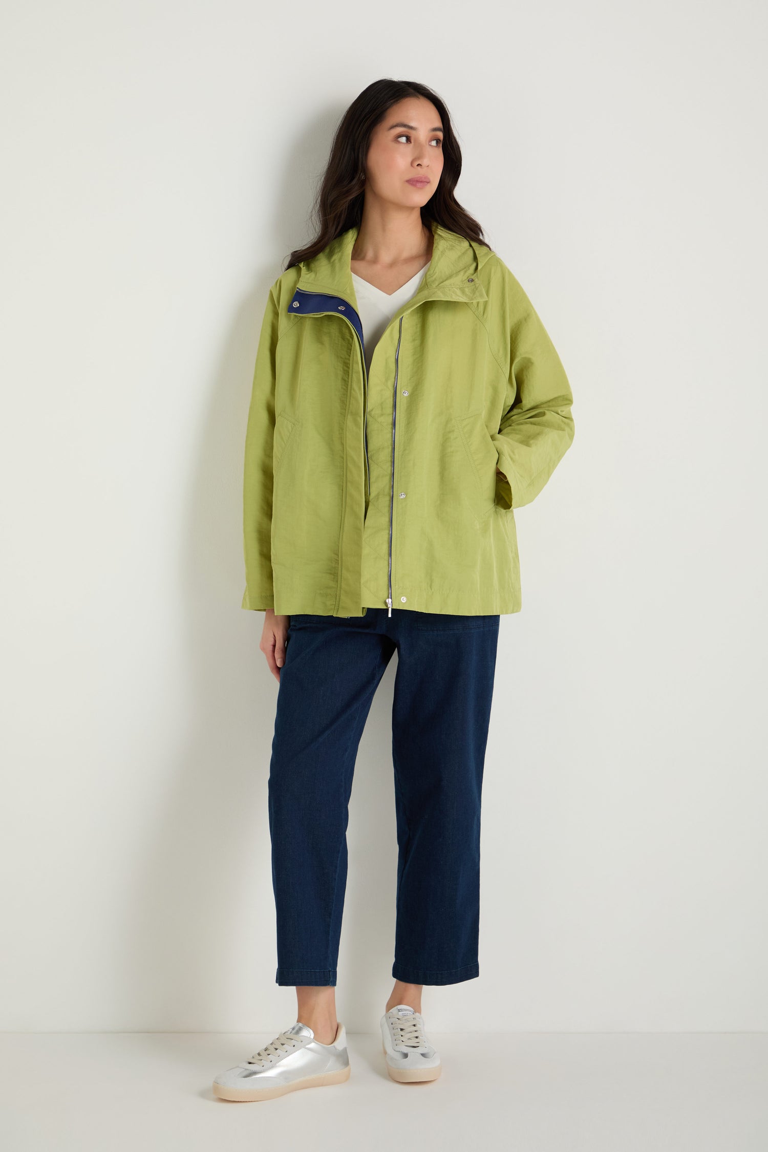 A woman stands against a white wall wearing a light green Showerproof Boxy Rain Jacket, a white top, dark blue pants, and white sneakers.