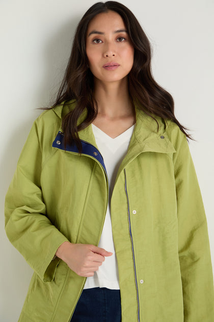 A woman with long dark hair wears the Showerproof Boxy Rain Jacket in light green over a white V-neck shirt, standing against a plain light background.