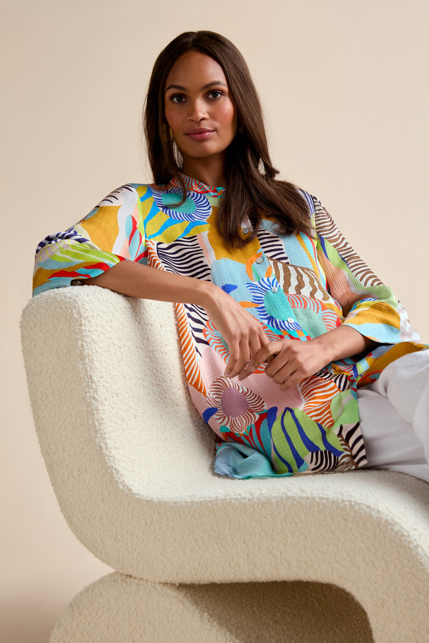 A woman sits on a modern cream boucle chair, wearing the Tropical Fantasy Shirt with abstract floral and zebra prints and relaxed fit, paired with white pants, against a neutral backdrop.