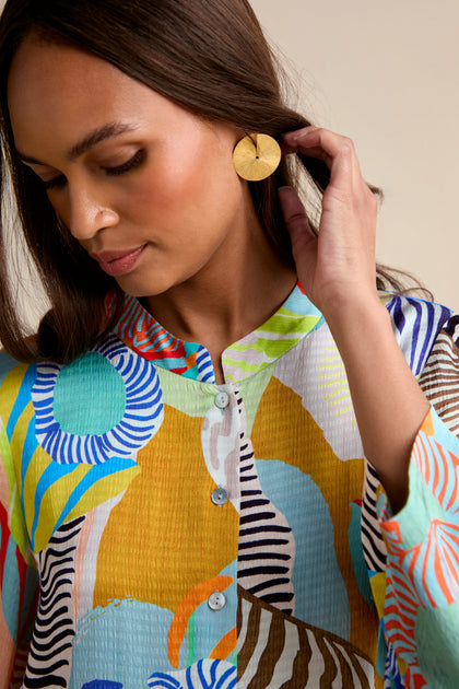 A woman wearing the vibrant Tropical Fantasy Shirt and large round gold earrings looks down while touching her hair.