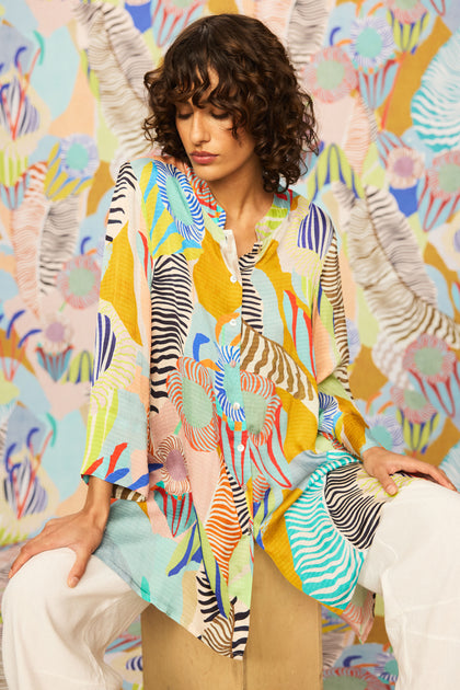 A woman with short curly hair wears the Tropical Fantasy Shirt, a relaxed-fit, textured viscose top with a vibrant tropical orchid print, paired with white pants and seated against a matching abstract multicolored backdrop.