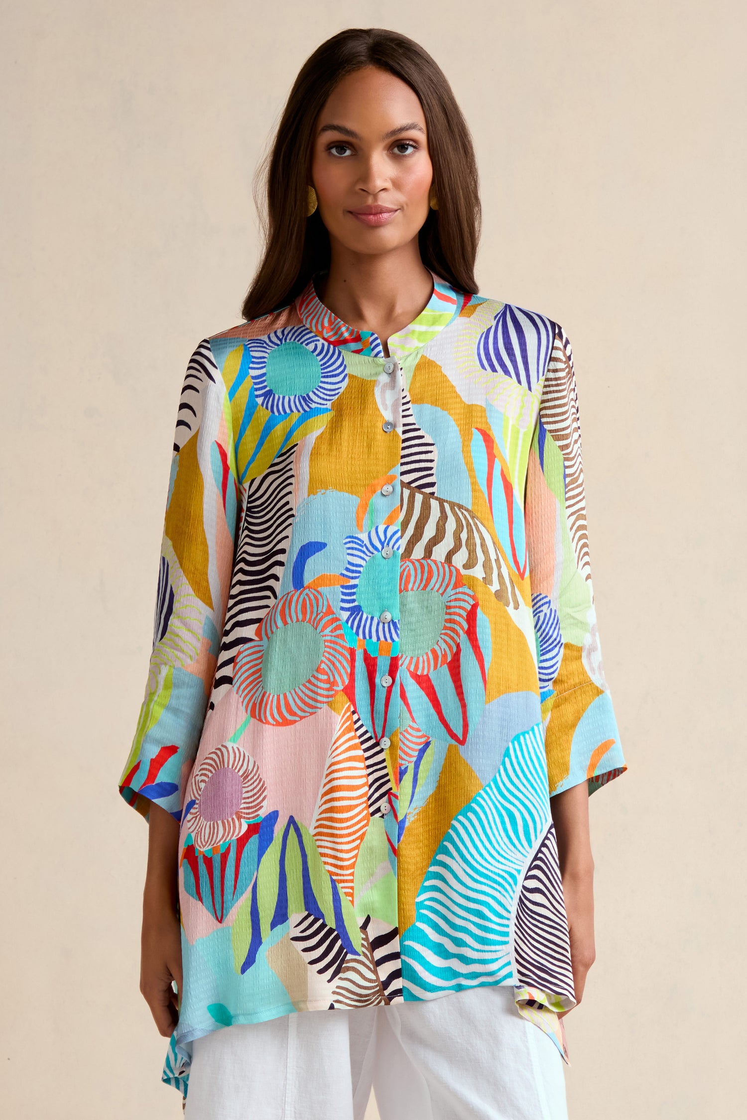A woman stands against a plain light background wearing the Tropical Fantasy Shirt, featuring a colorful tropical print and long sleeves, paired with white pants.