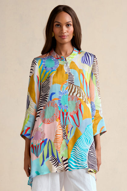 A woman stands against a plain light background wearing the Tropical Fantasy Shirt, featuring a colorful tropical print and long sleeves, paired with white pants.