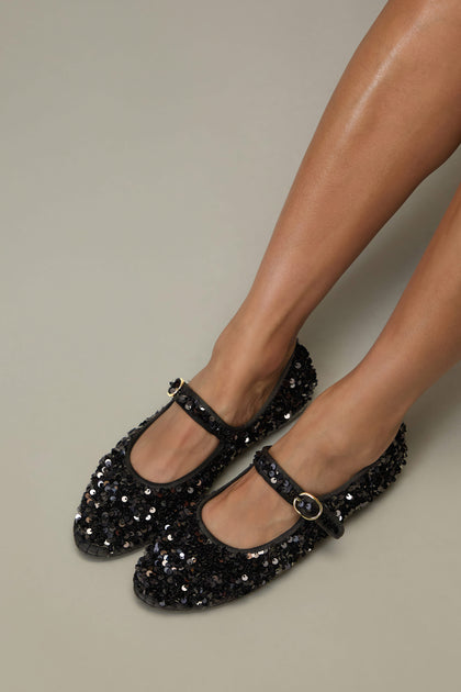 Someone is shown from the knees down wearing Sequin Mary Jane Velvet Shoes—black, handmade in Spain with sequin detail, a strap, and buckle—set against a neutral background.