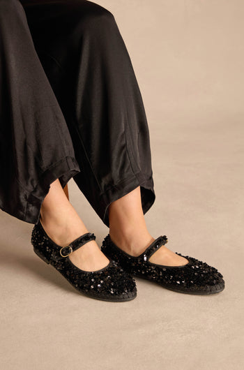 Black sequined Sequin Mary Jane Shoes with an ankle strap worn with black trousers.