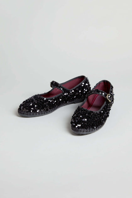 Sequin Mary Jane Shoes, handmade in Spain, feature a black sequin-textured surface, a secure strap with buckle, and a dark red interior lining, displayed on a light background.