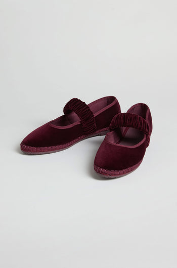 The Mafalda Wine Velvet Shoes, handcrafted in Spain, feature thick ruched straps and flat soles. This maroon Mary Jane pair is displayed on a plain white background.