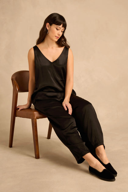 A woman in a black sleeveless top and trousers sits on a wooden chair wearing handmade Velvet Slipper Shoes.