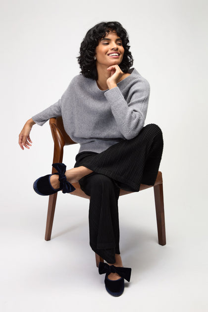 A person with short curly hair sits on a wooden chair, wearing a gray sweater, black pleated pants, and navy Lyra Bow velvet shoes from Flabelus, smiling and resting an arm on the chair back.