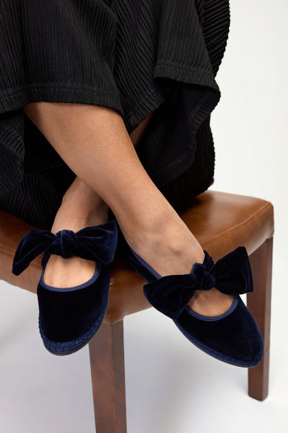 A person wearing a long black pleated skirt and Navy Lyra Bow Velvet Shoes, seated on a brown wooden chair.