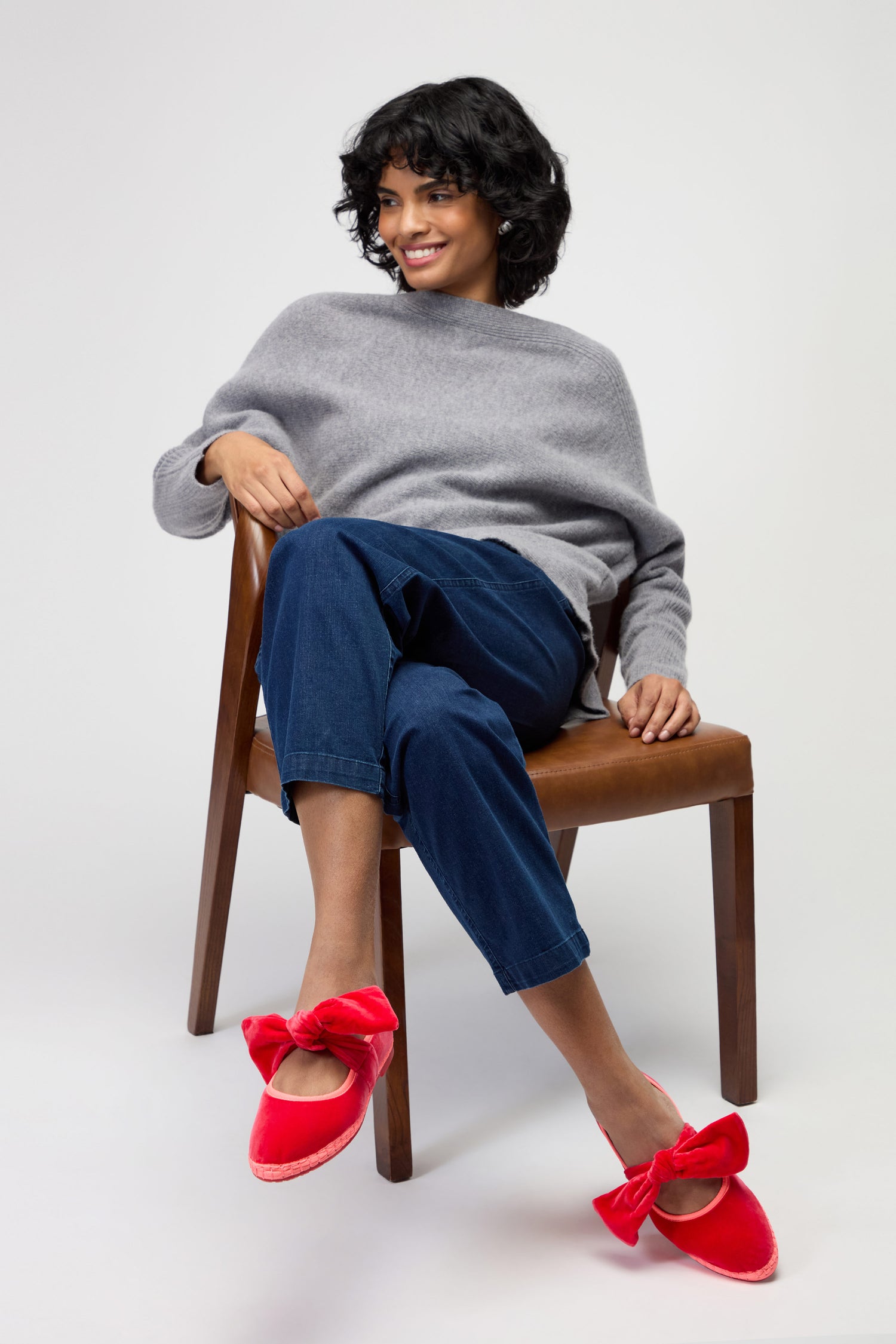 A woman sits on a wooden chair wearing a gray sweater, blue pants, and Strawberry Pink Lyra Bow Velvet Shoes against a plain white background.