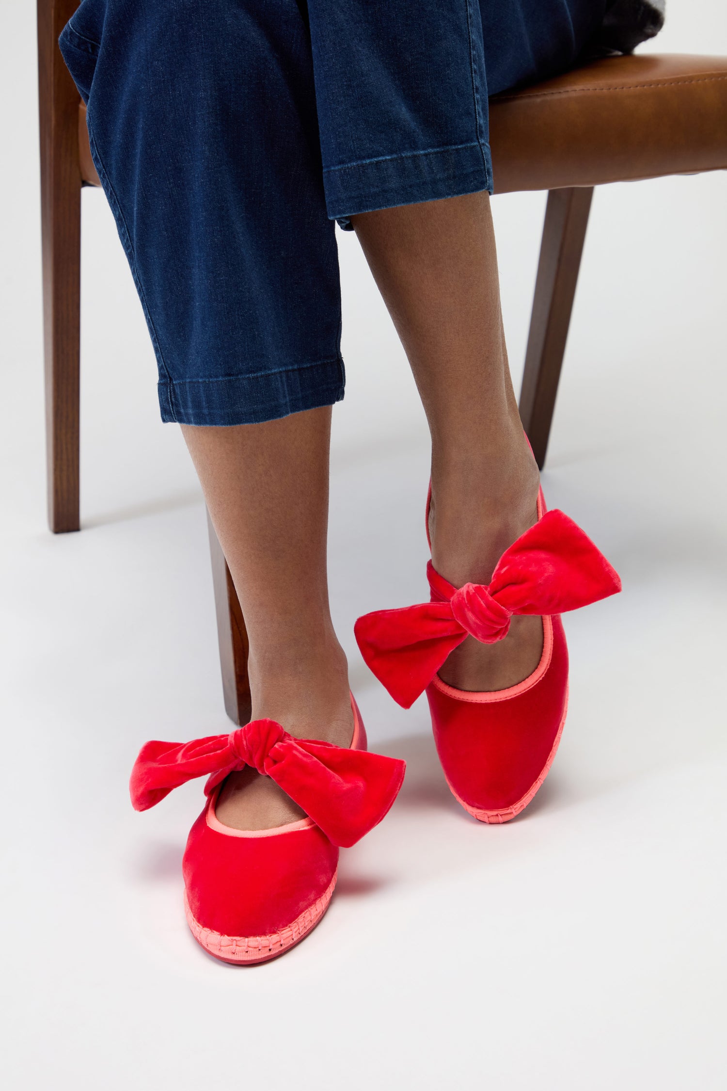 A person seated, wearing dark blue pants and Strawberry Pink Lyra Bow Velvet Shoes, their feet resting on the floor.
