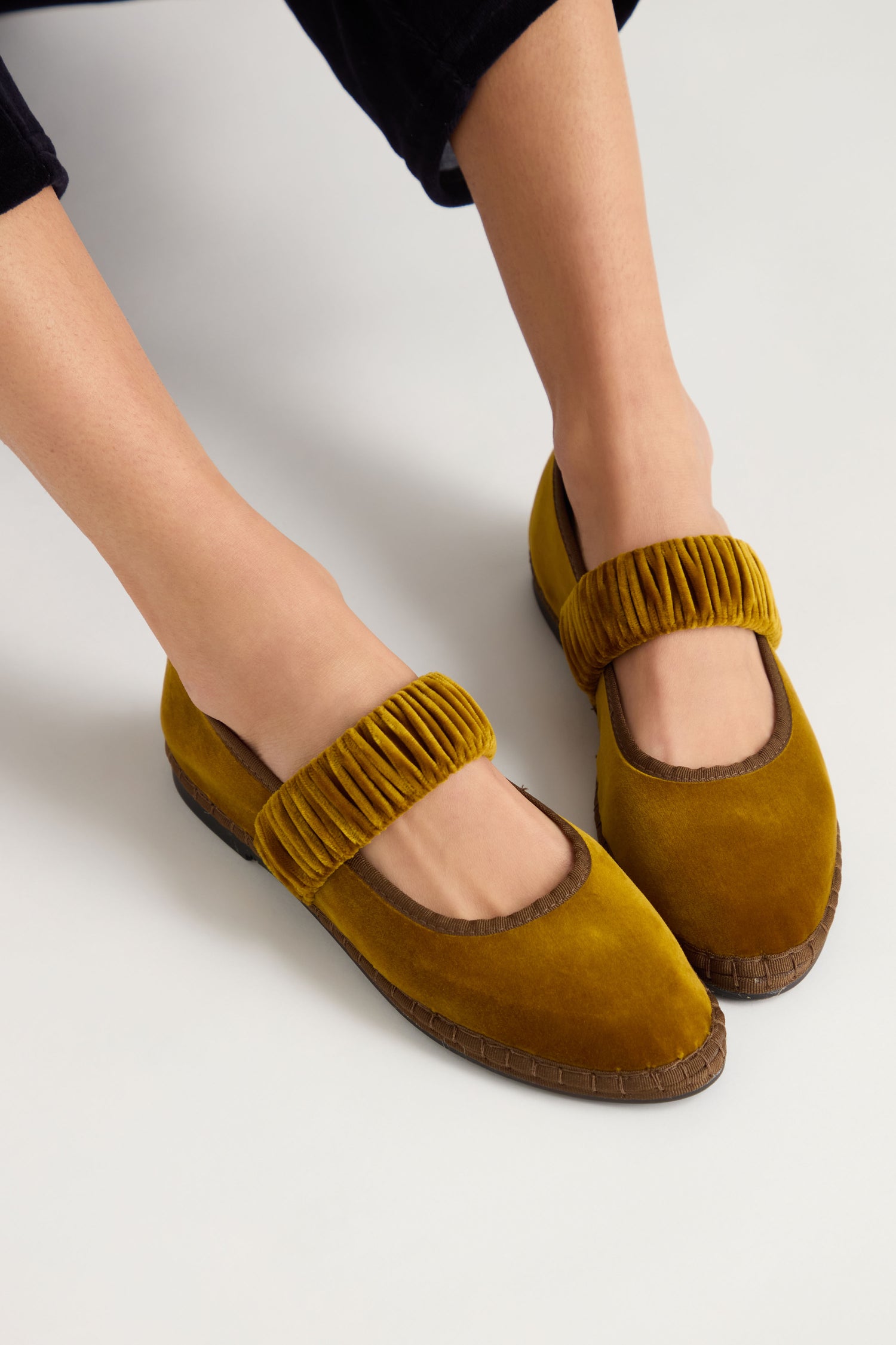 A person is shown from above wearing Mafalda Ochre Velvet Shoes—mustard yellow Mary Jane flats with pleated straps and brown soles, handmade in Spain from recycled rubber.