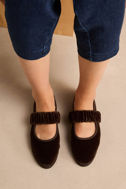 Wearing cropped blue jeans, the person stands on a beige floor in Mafalda Truffle Velvet Shoes—dark brown Mary Janes with textured straps and a recycled rubber sole.