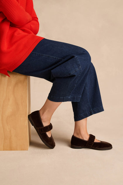 Wearing the Mafalda Truffle Velvet Shoes—featuring a ruffled strap detail and recycled rubber sole—a person in a red sweater and wide-leg blue jeans sits on a wooden bench.