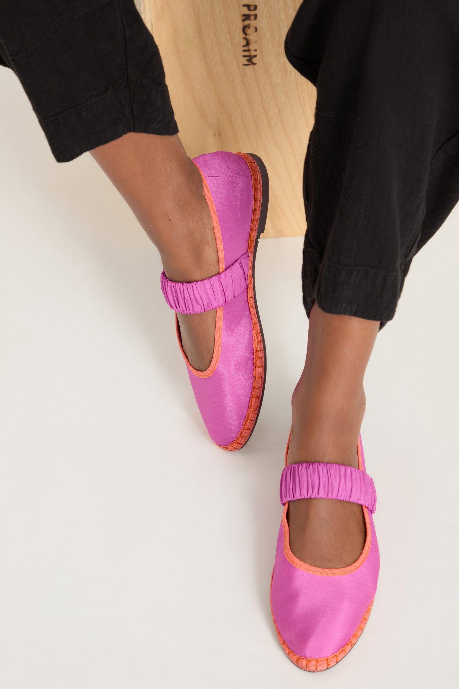 A person wearing black pants and Mafalda Silk Shoes—bright pink Mary Jane-style shoes with orange trim, handcrafted in Spain—sits on a light wooden stool.