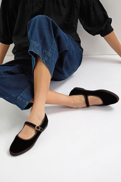 A person sits on a white floor against a neutral background, wearing blue denim pants and Black Mary Jane Velvet Shoes featuring a gold buckle and crafted with traditional Spanish shoemaking techniques.