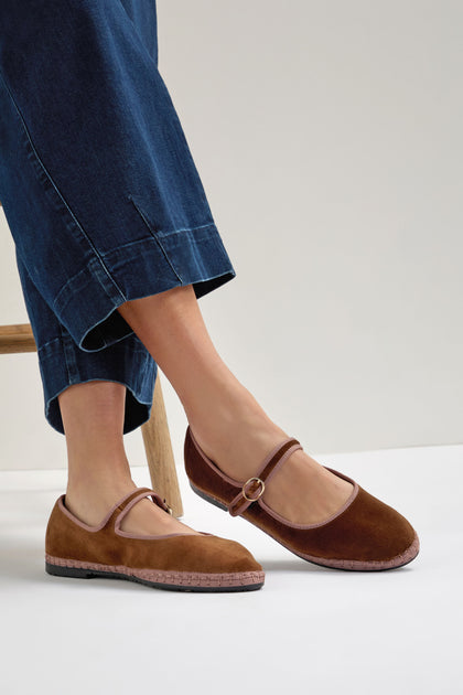 A person in denim pants is seated, showcasing Brown Mary Jane Velvet Shoes with a strap and buckle for a vintage touch.