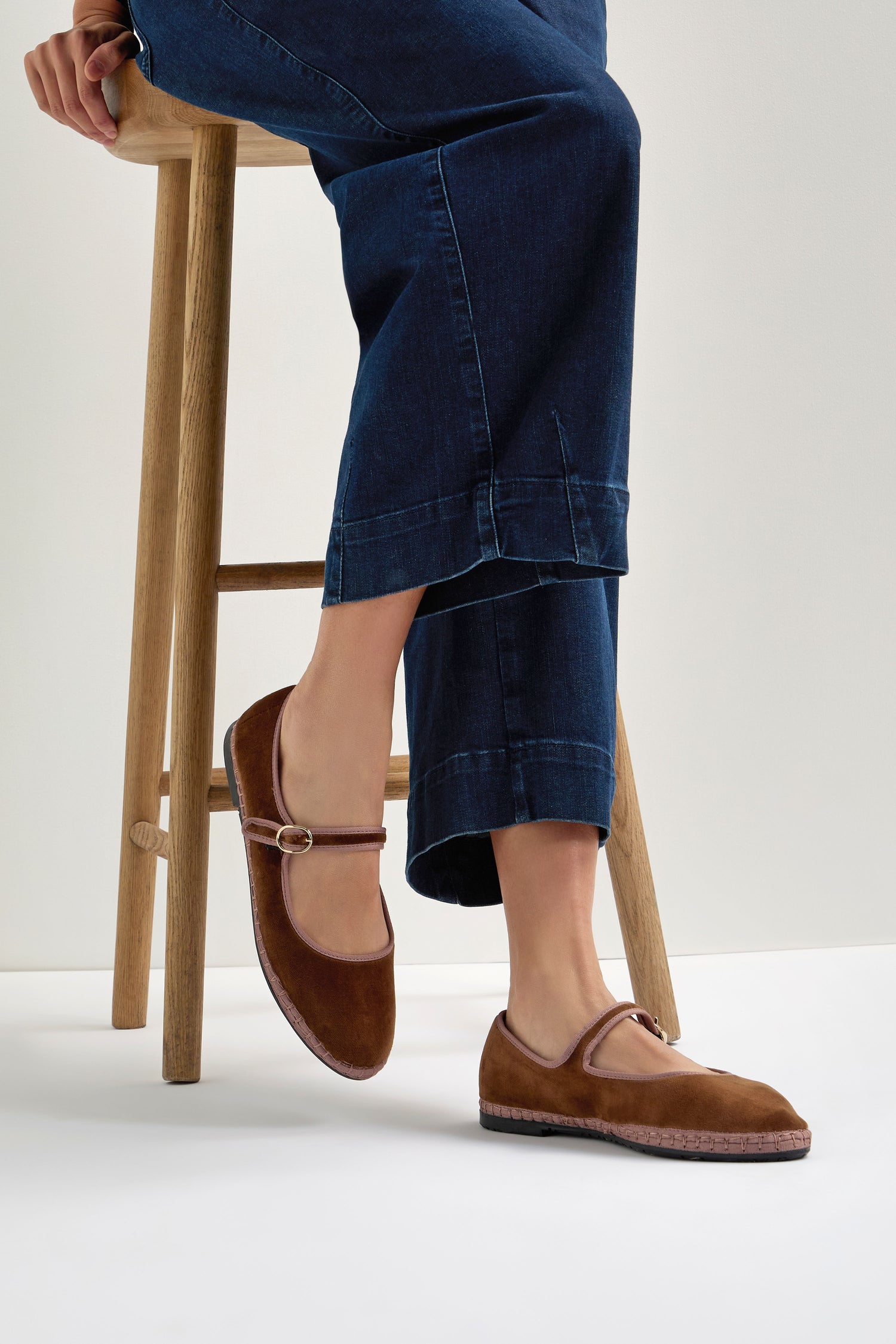 A person wearing dark blue jeans sits on a wooden stool, showcasing Brown Mary Jane Velvet Shoes featuring a small buckle that adds vintage charm to their classic look.