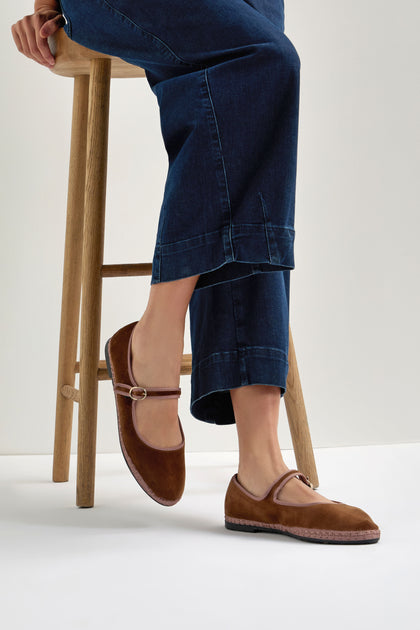 A person wearing dark blue jeans sits on a wooden stool, showcasing Brown Mary Jane Velvet Shoes featuring a small buckle that adds vintage charm to their classic look.