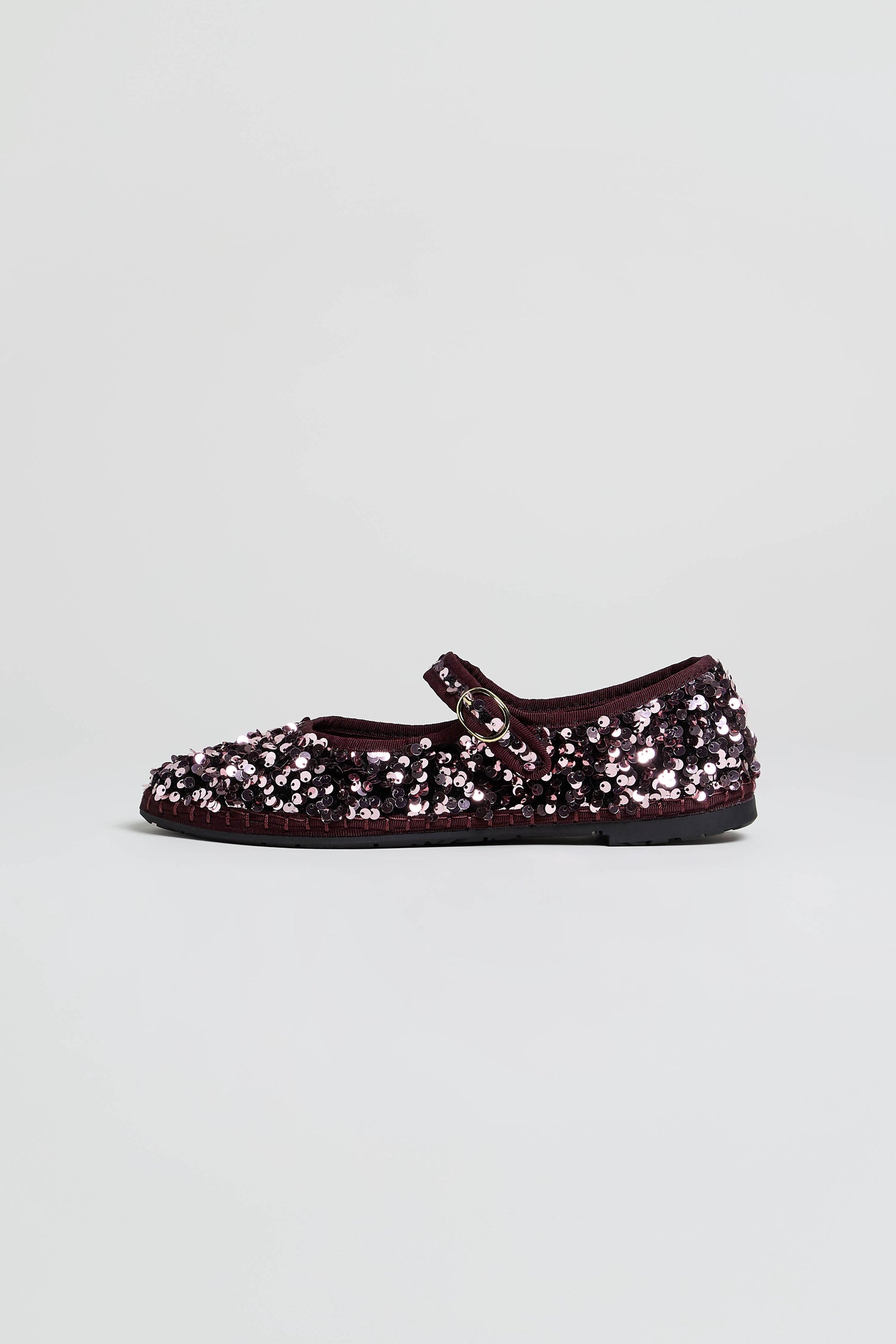 A single Sequin Mary Jane Shoe in maroon, handcrafted in Spain and adorned with sparkling sequins, showcases a side buckle strap and the refined look of a velvet flat, set against a simple light background.