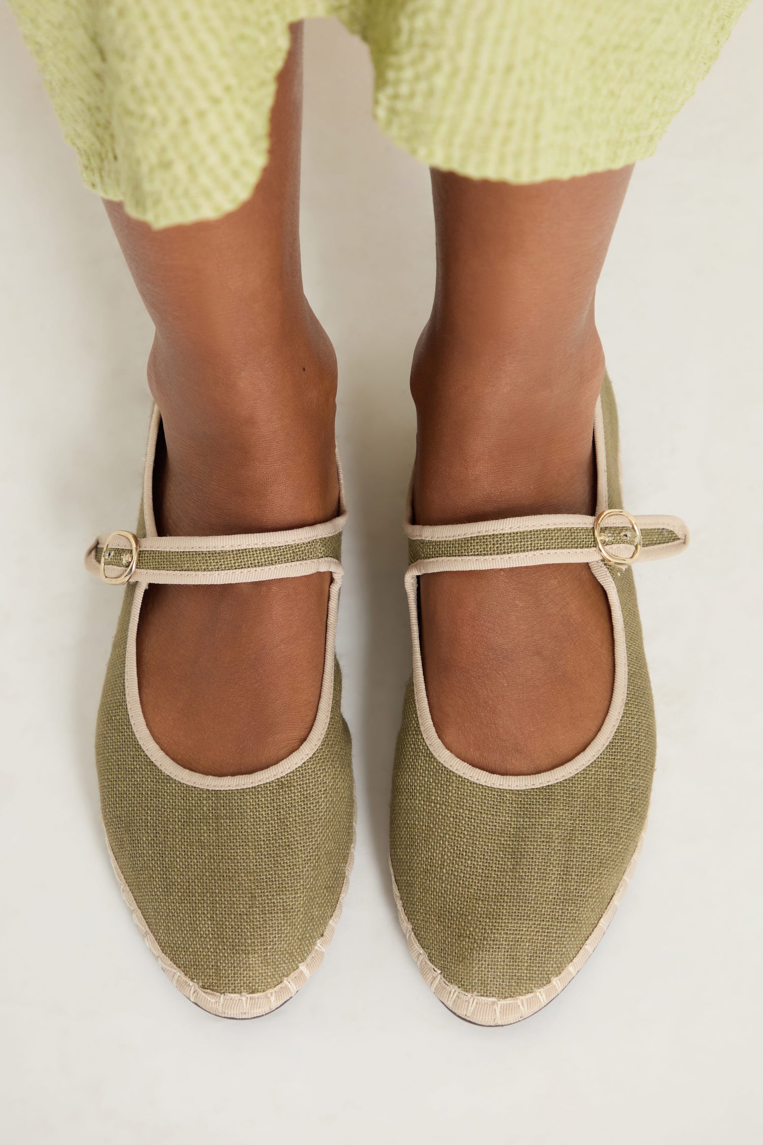 A person stands on a white surface wearing a light green textured skirt and Mary Jane Linen Shoes in olive green with beige trim, buckle, recycled rubber sole, and organic cotton lining.