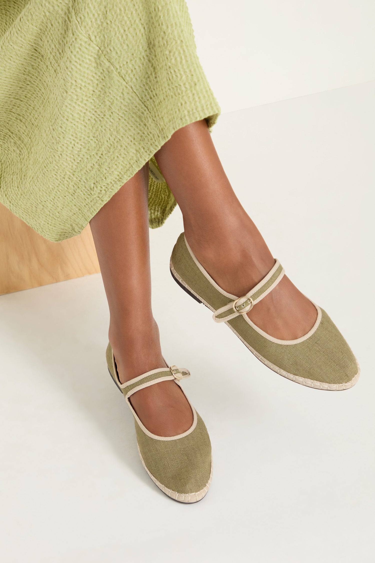 A person wearing a light green textured skirt and the Mary Jane Linen Shoes with beige trim and recycled rubber sole sits on a wooden bench against a white background.