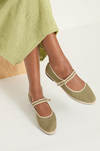 A person wearing a light green textured skirt and the Mary Jane Linen Shoes with beige trim and recycled rubber sole sits on a wooden bench against a white background.