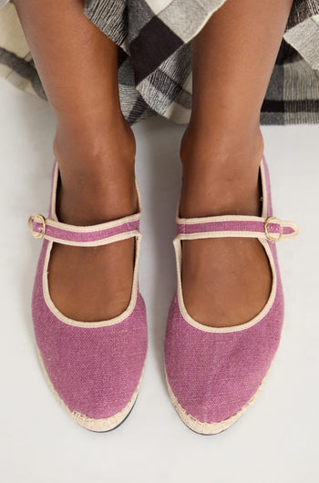 A person wearing Mary Jane Linen Shoes in mauve with beige trim and buckle straps, organic cotton lining, and recycled rubber sole stands on a white surface, partially covered by a plaid skirt.