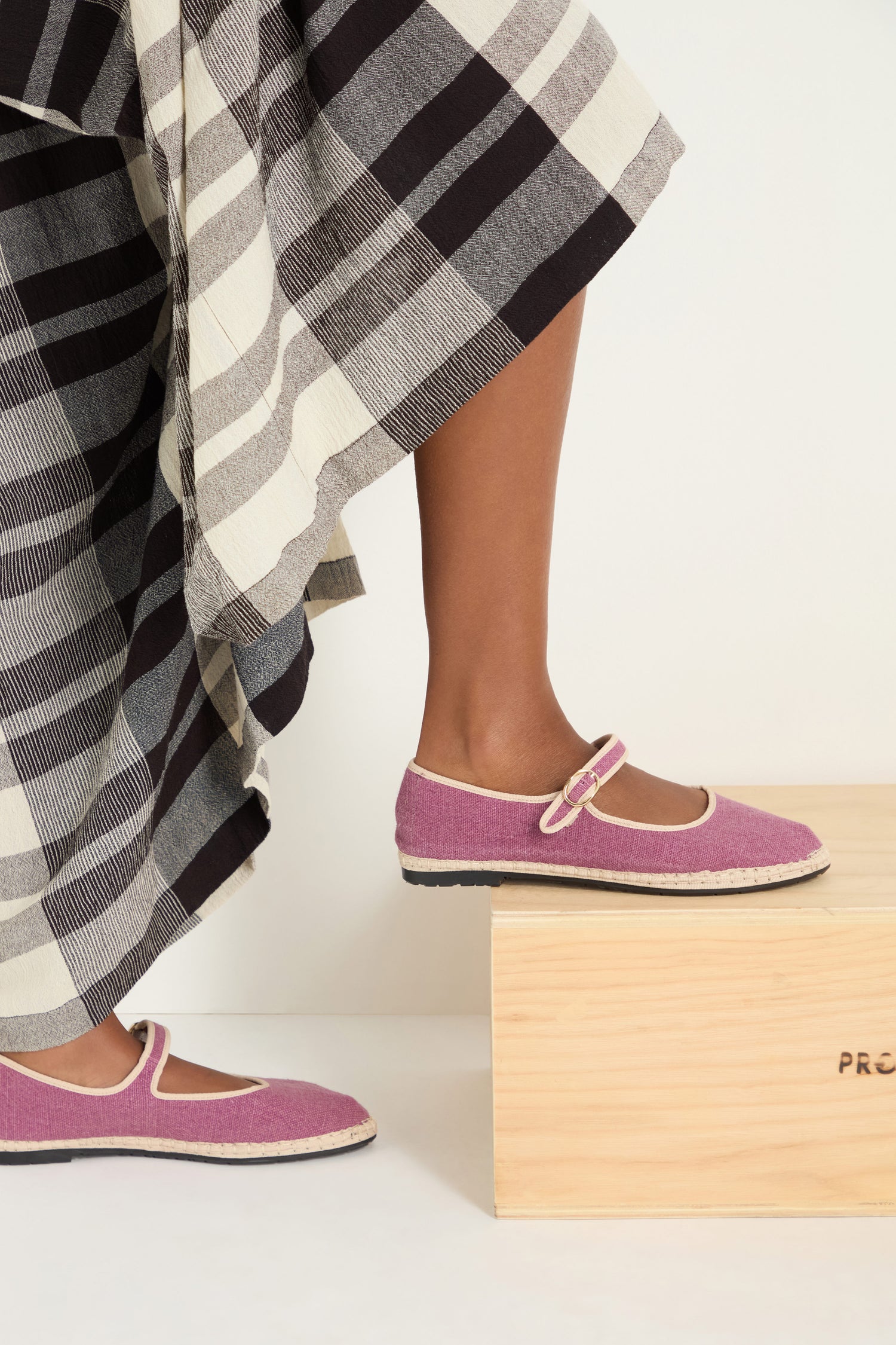 A person wearing a long, black-and-white striped skirt and Mary Jane Linen Shoes made with an organic cotton lining and recycled rubber sole stands with one foot on a wooden box.