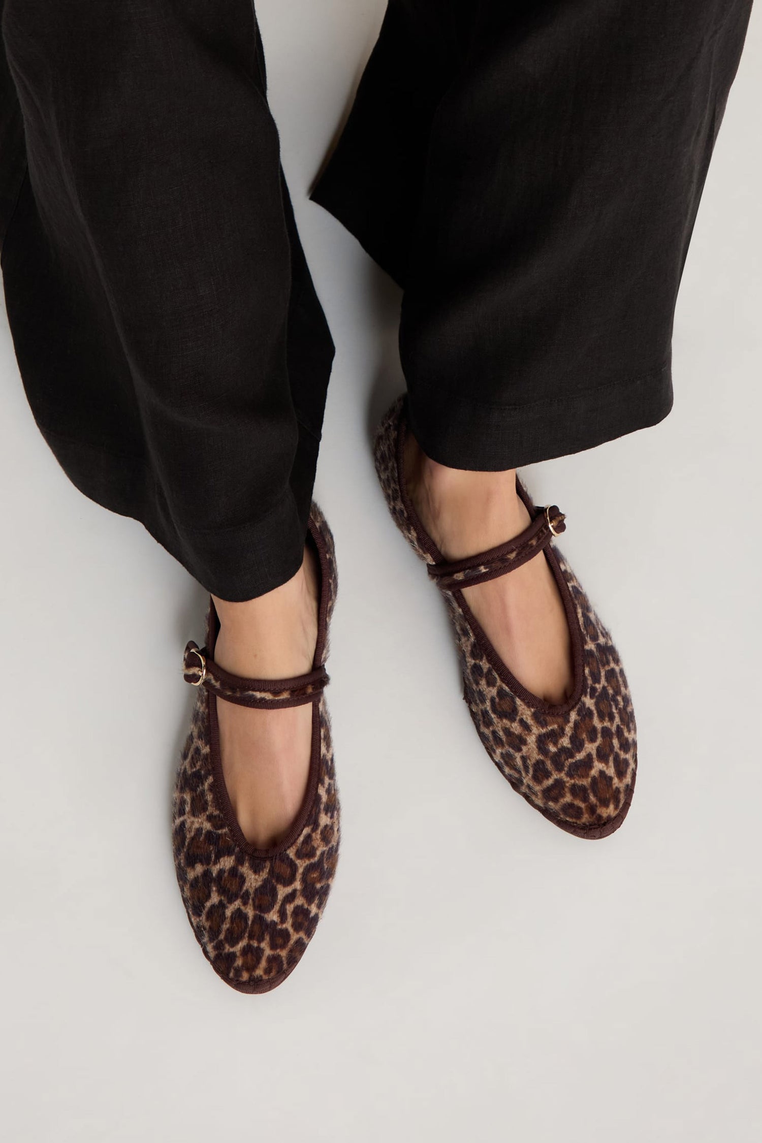 A person stands on a light surface wearing black pants and Cheetah Mary Jane Shoes—handmade in Spain with ankle straps and a recycled bicycle tyre sole.