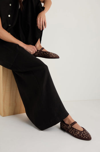 A person sits on a wooden block wearing wide-leg black pants, a black top, and Cheetah Mary Jane Shoes—handmade in Spain with recycled bicycle tyre soles. One foot rests on the floor while the other leg is bent.