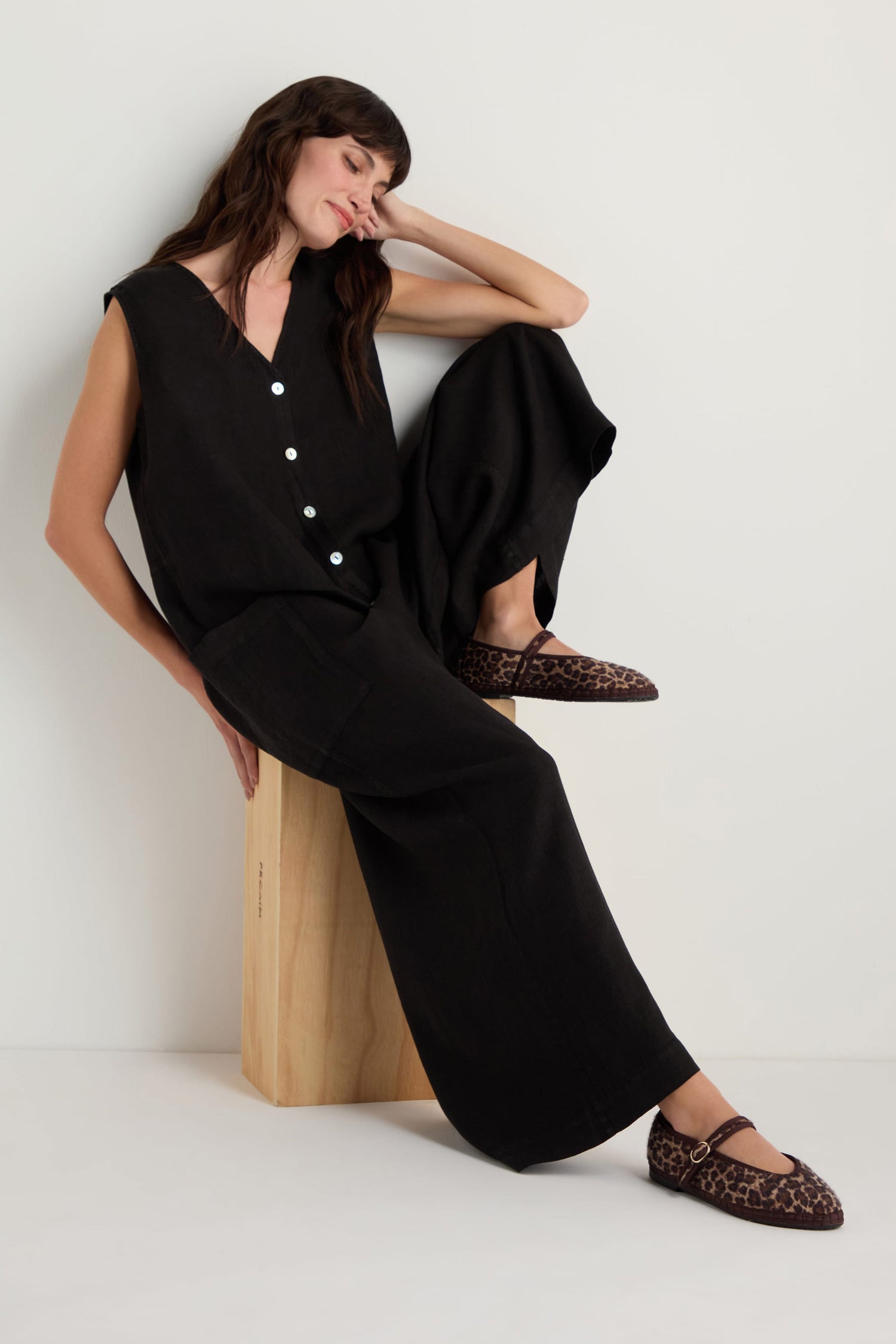 A woman in a sleeveless black jumpsuit sits on a wooden block, resting her head on her hand, wearing Cheetah Mary Jane Shoes with recycled bicycle tyre soles, handmade in Spain, against a plain white background.