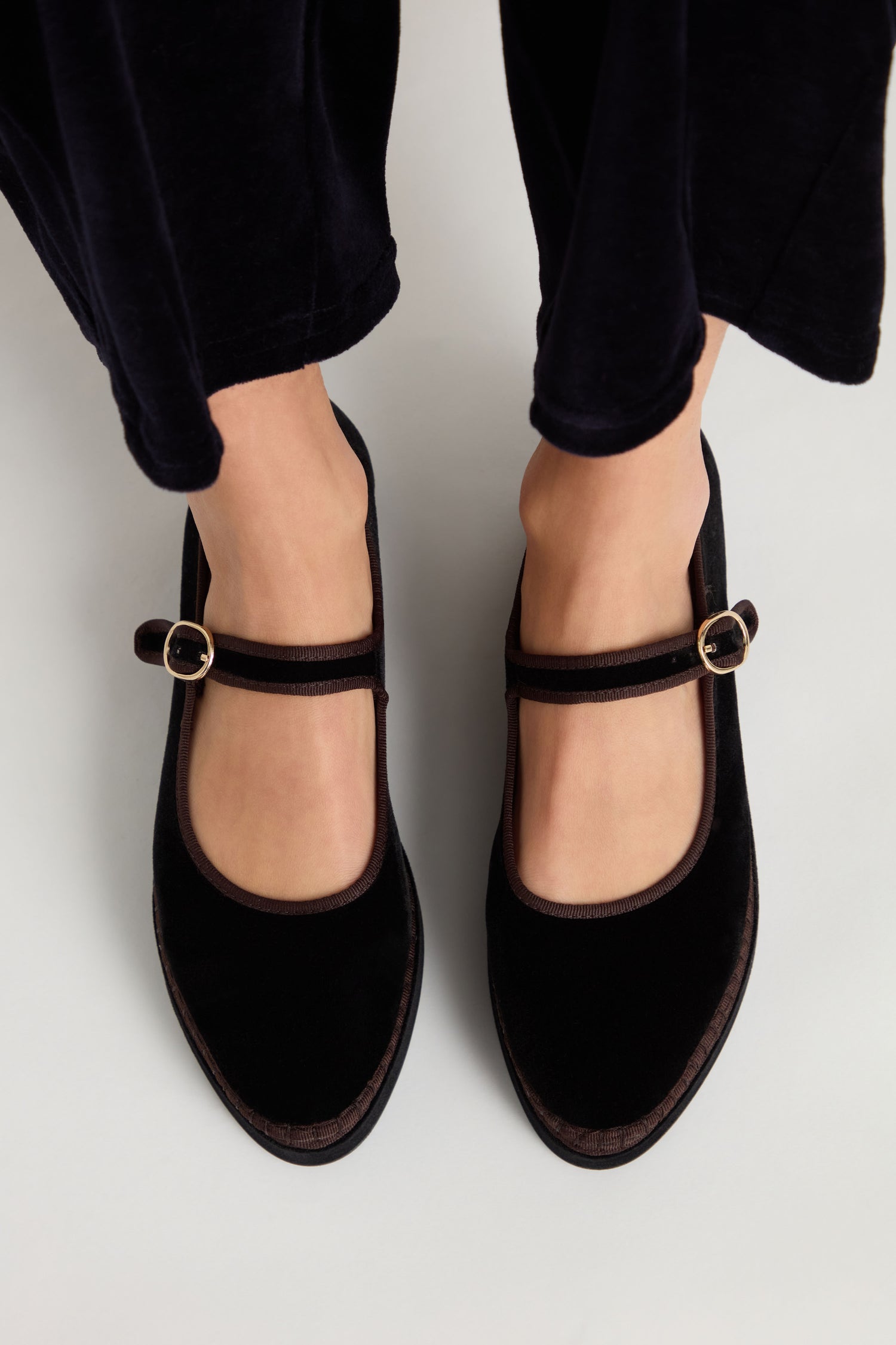 A person is wearing Black Velvet Platform Mary Jane Shoes with buckle straps, standing on a light-colored surface. The handmade shoes are styled with dark pants.